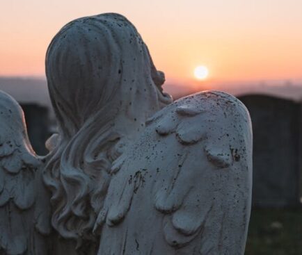A contemplative angel statue in a cemetery, facing a serene sunset sky.