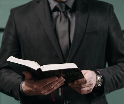 Elegant man in a suit holding an open book with flowers in the foreground, conveying solemnity.